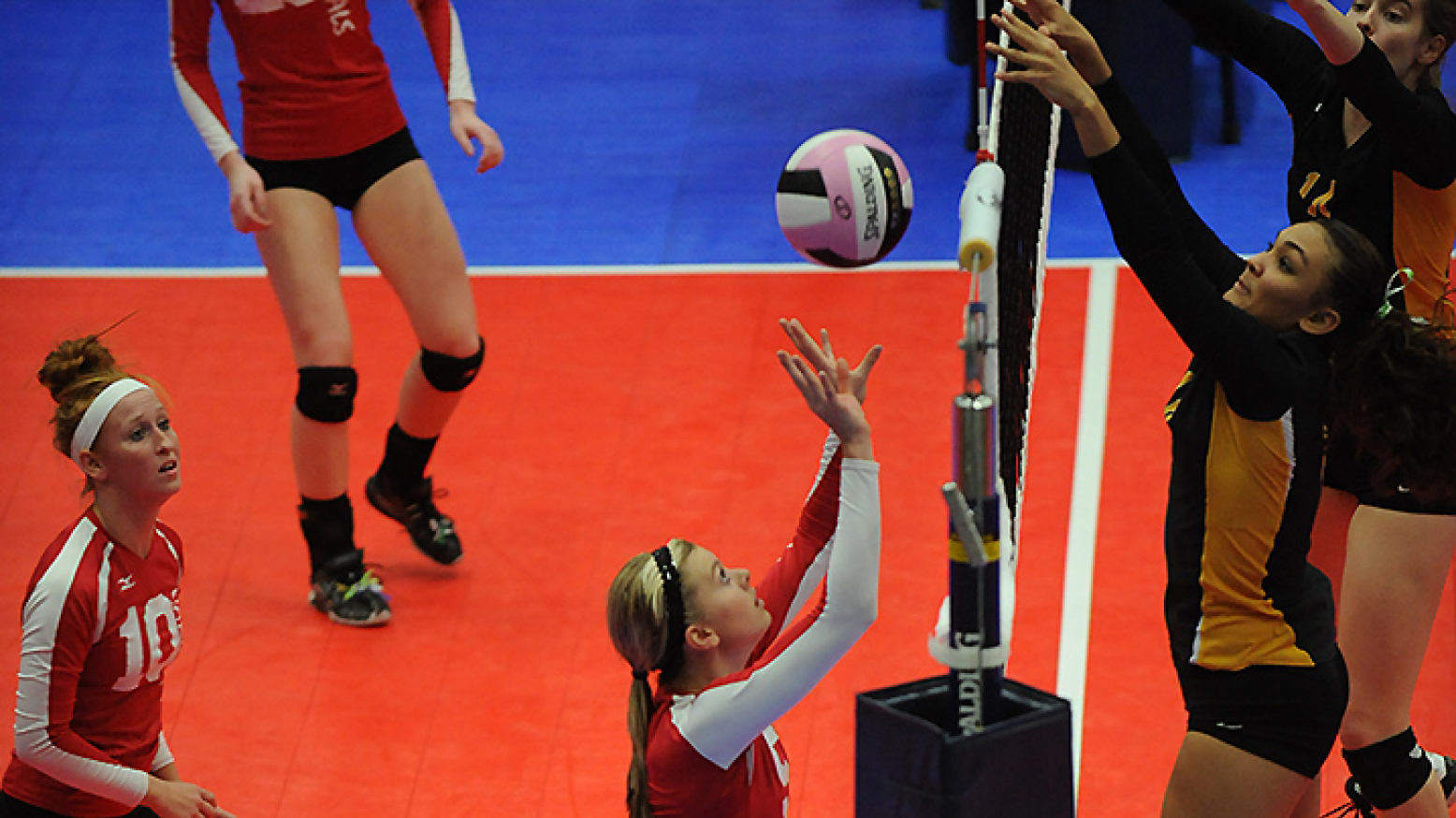 Female volleyball players on a bright red and blue court. A player at the center of the image is about to hit the ball upward while multiple opponents have their hands up at the net, preparing to block the ball. 