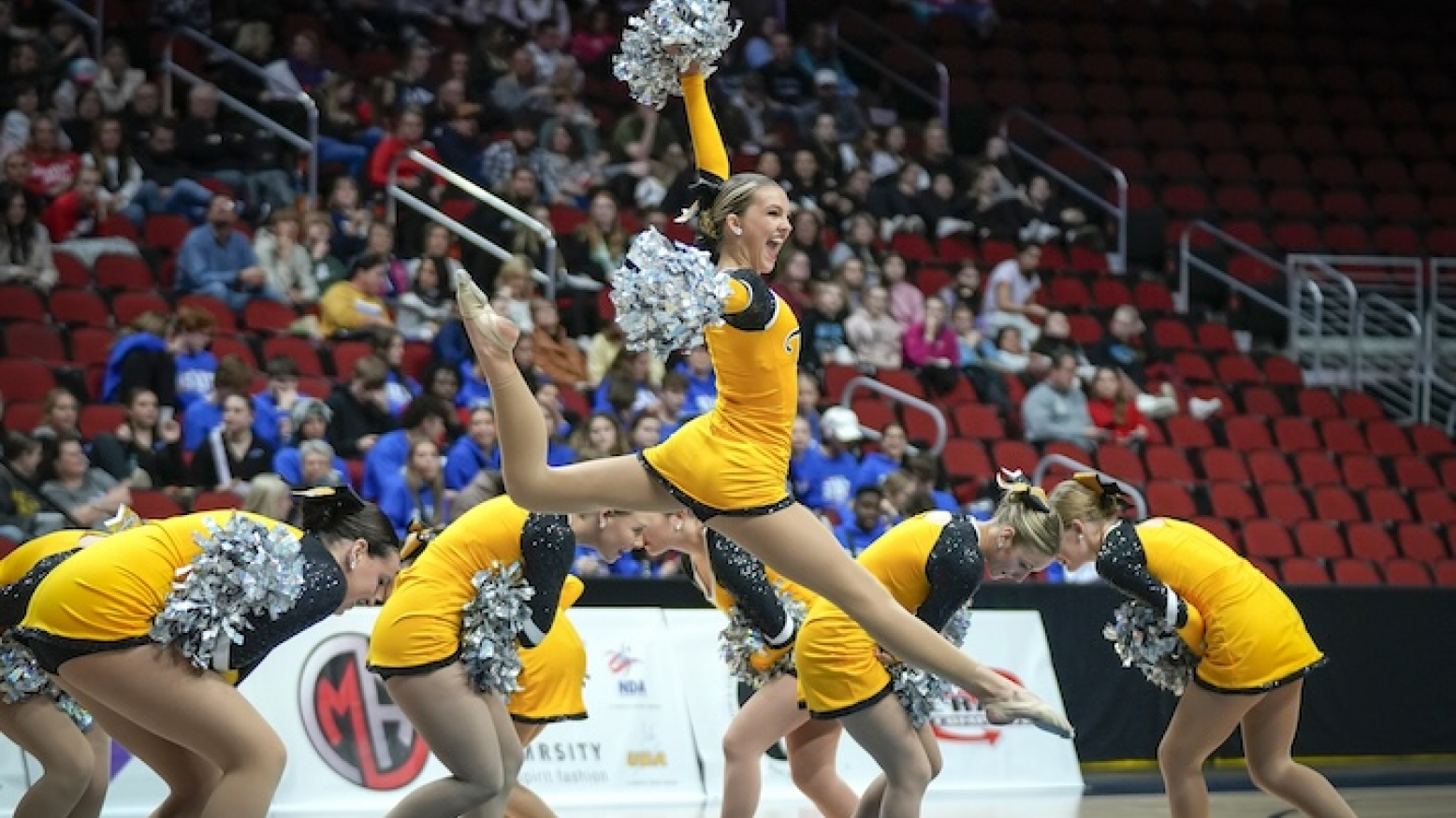 Female dancers performing in yellow uniforms with silver pom poms. The dancer in the center foreground is leaping in the air with her arms raised.