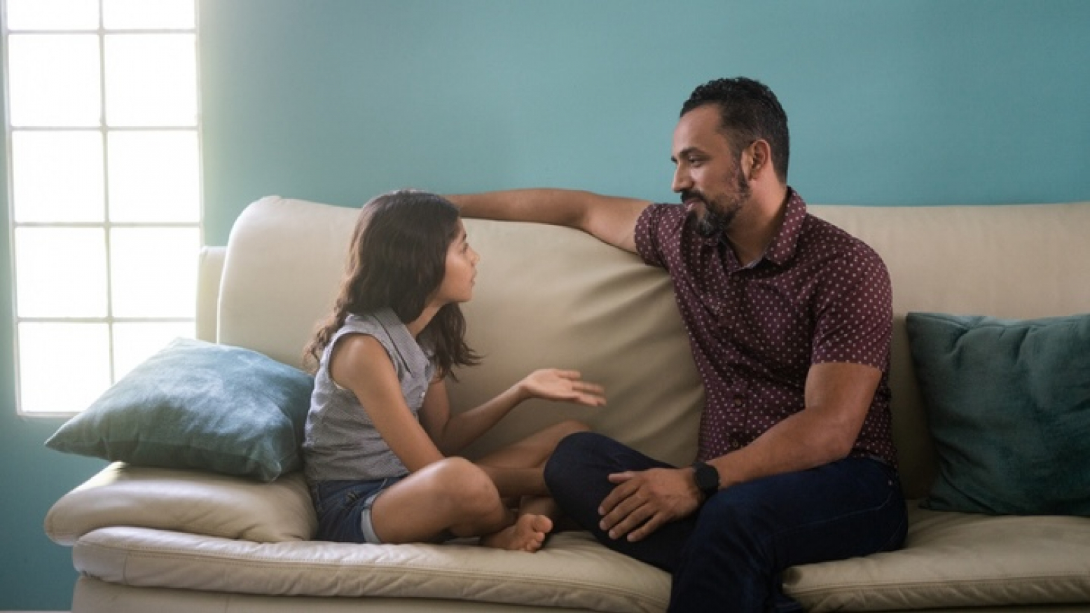 A Latinx father and his young daughter sitting on the sofa at home talking face to face.