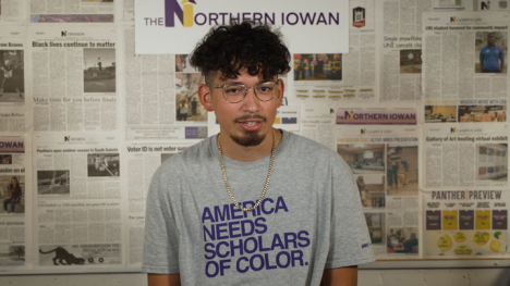 Nixson Benitez stands in front of a bulletin board covered in newspapers. His shirt reads America Needs Scholars of Color.
