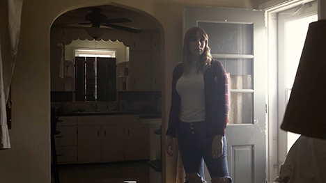 A woman stands at the entryway of a house with the outside light pouring into a dimly lit room.