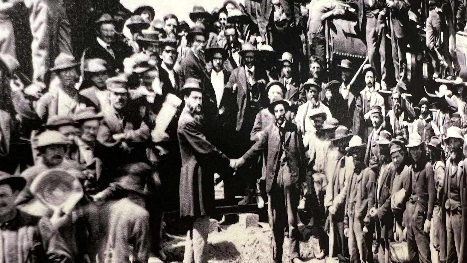A historic, black and white photo of people shaking hands in front of a crowd at the completion of the Transcontinental Railroad.