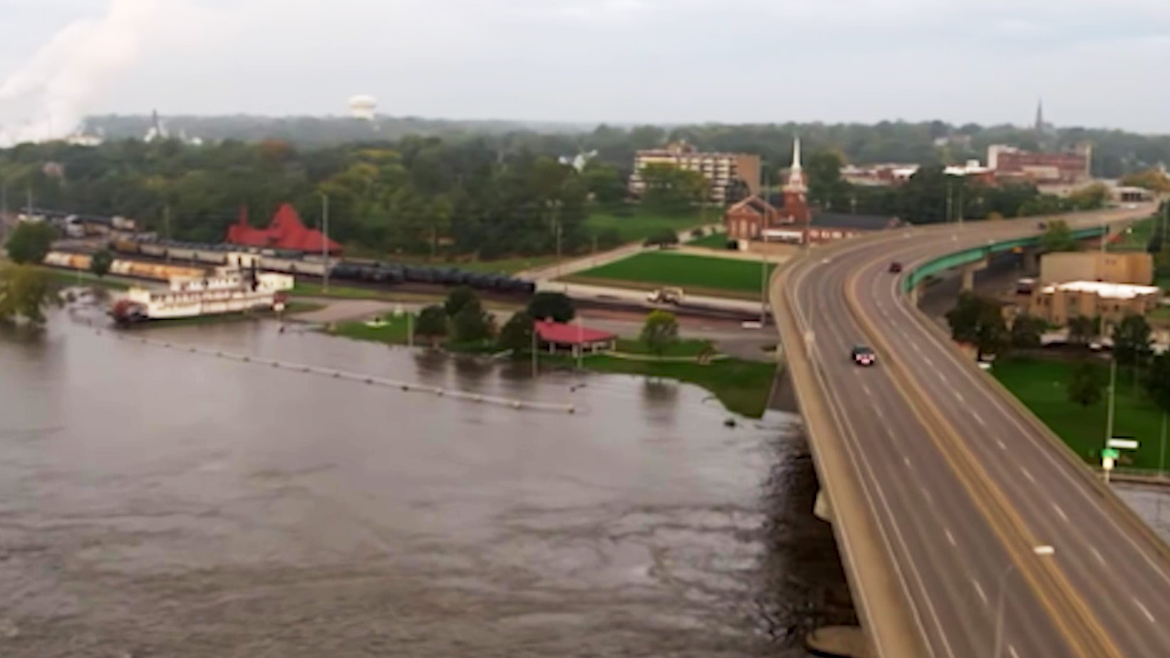 A road bridge over a large river, with a train running along tracks that are parallel to the river.