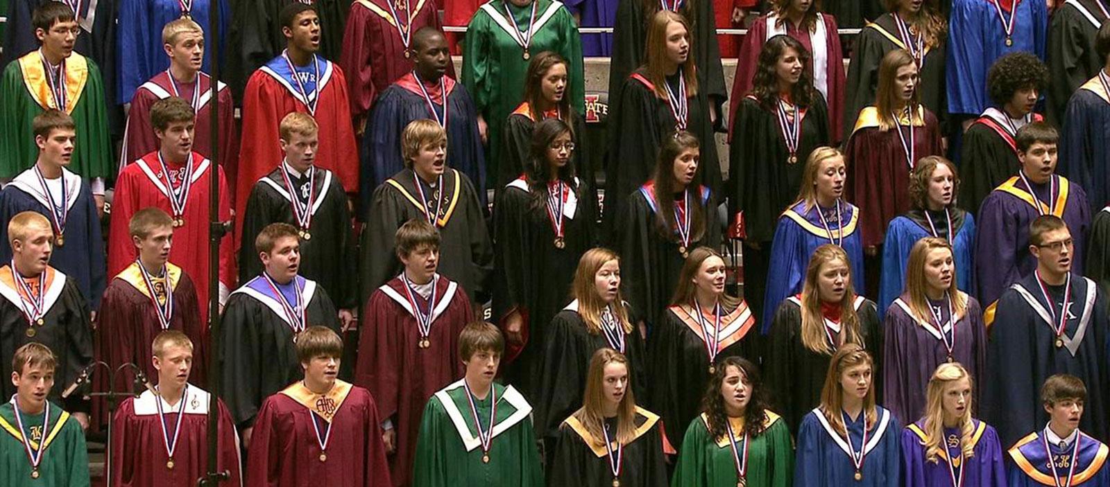 Rows of high school students in various colors of robes are singing.