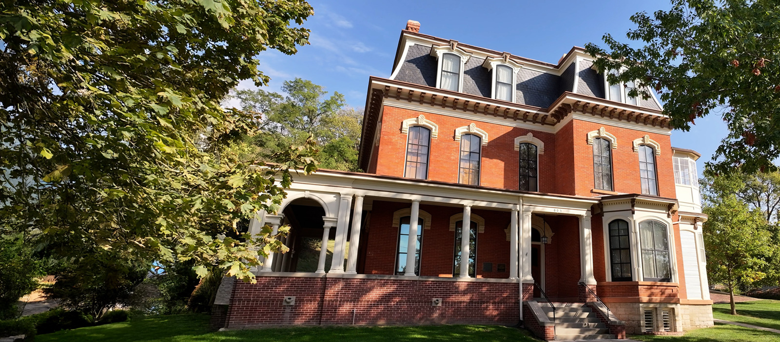 A Victorian and Italianate 3 story brick house with a mansard roof sits atop a hill.