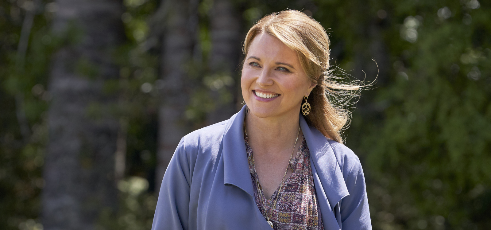 Lucy Lawless as Alexa Crowe smiling outdoors, wearing a light blue coat and holding a smartphone.