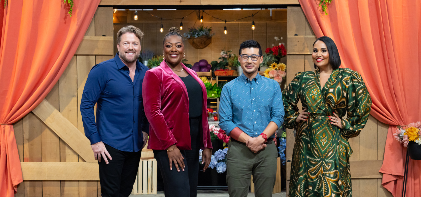 The three judges and host posing in front of a wooden backdrop with flowers and orange curtains.