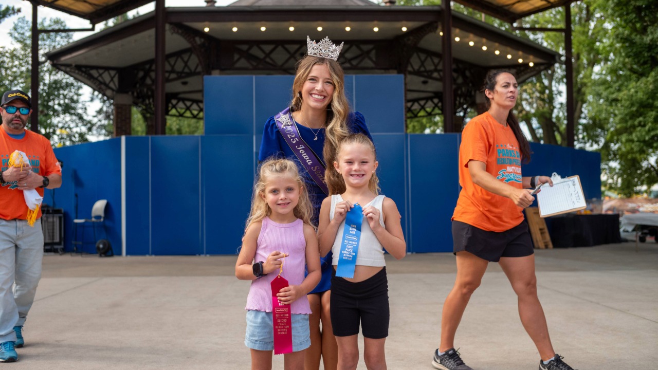 Fair Queen standing with a couple of little kids with ribbons for participating in the pie eating contest