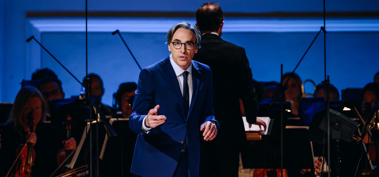 A man in a suit standing on stage in front of an orchestra