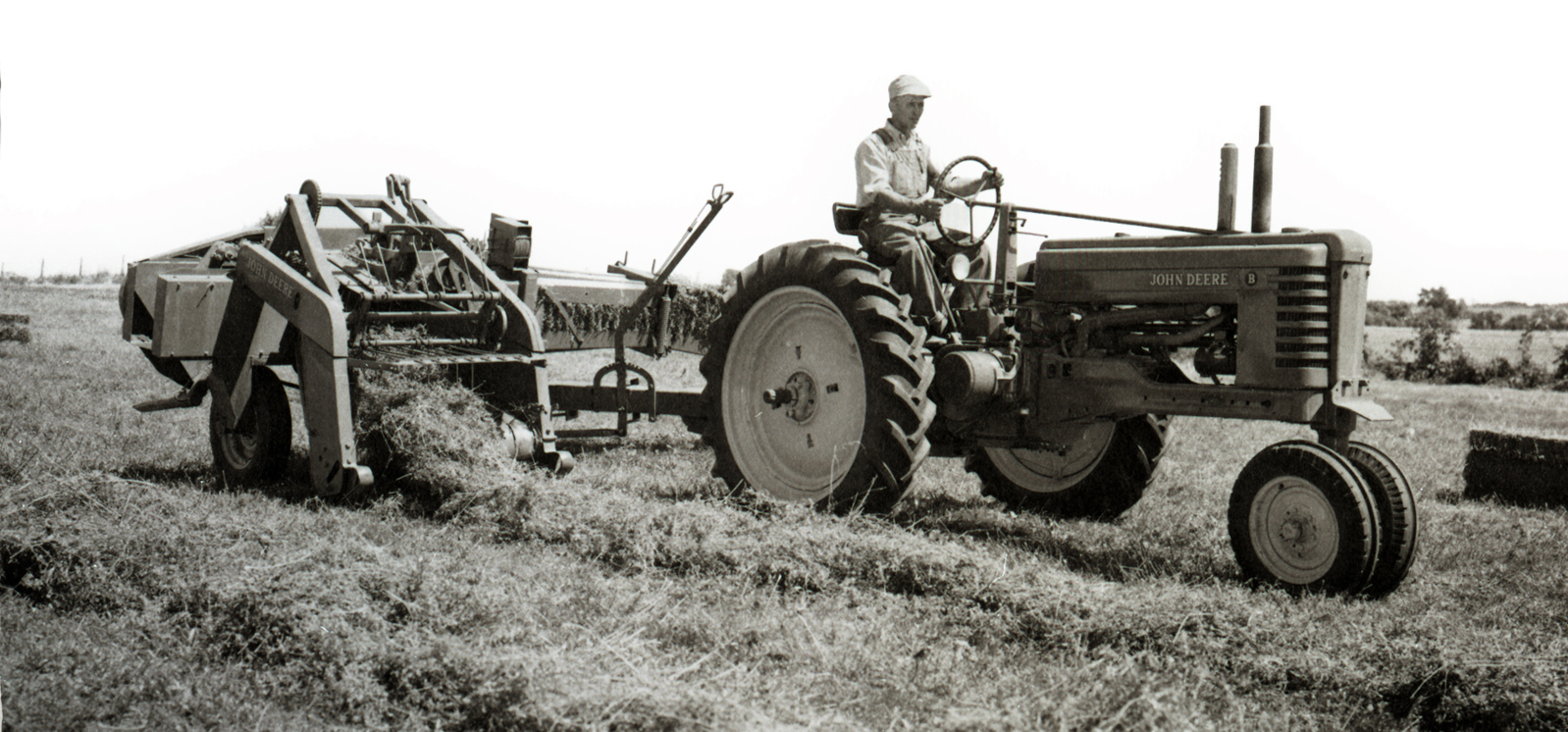 A person driving a John Deere tractor with attached hay baler in a field.