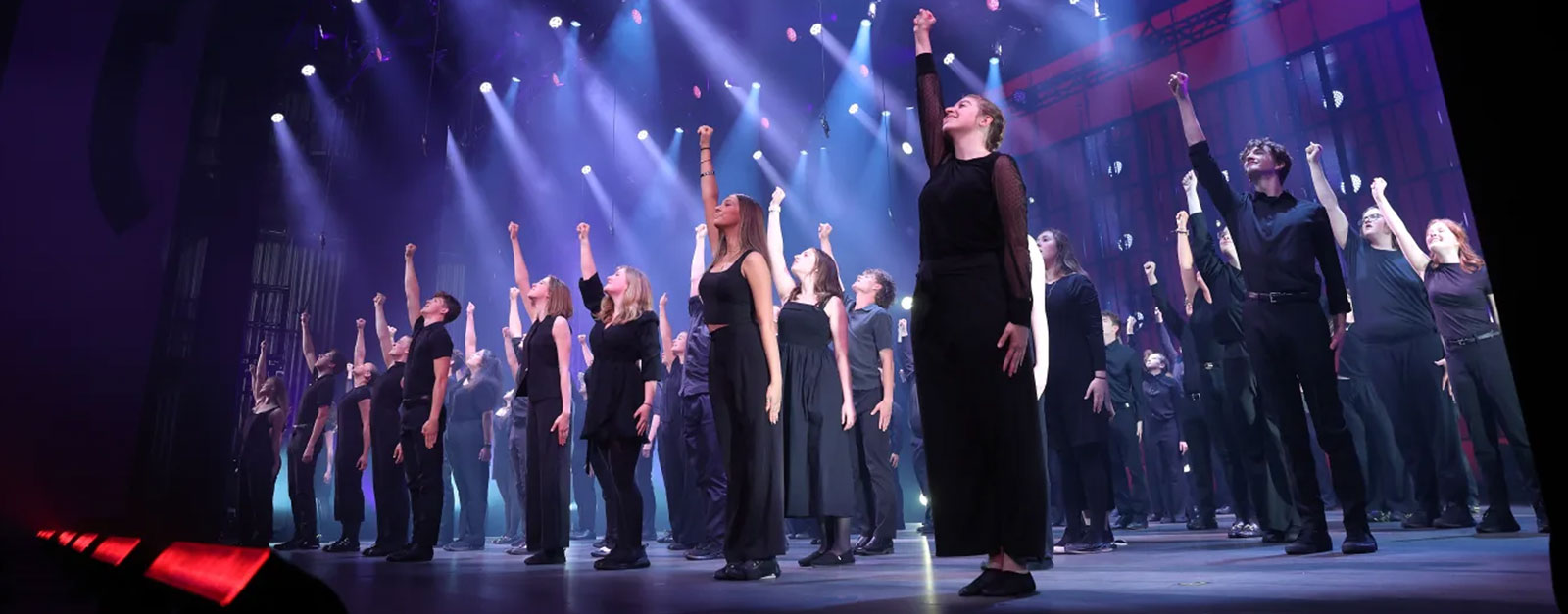 A stage is filled with teens dressed in black, each with an arm risen in the air, with spotlights shining down on them.