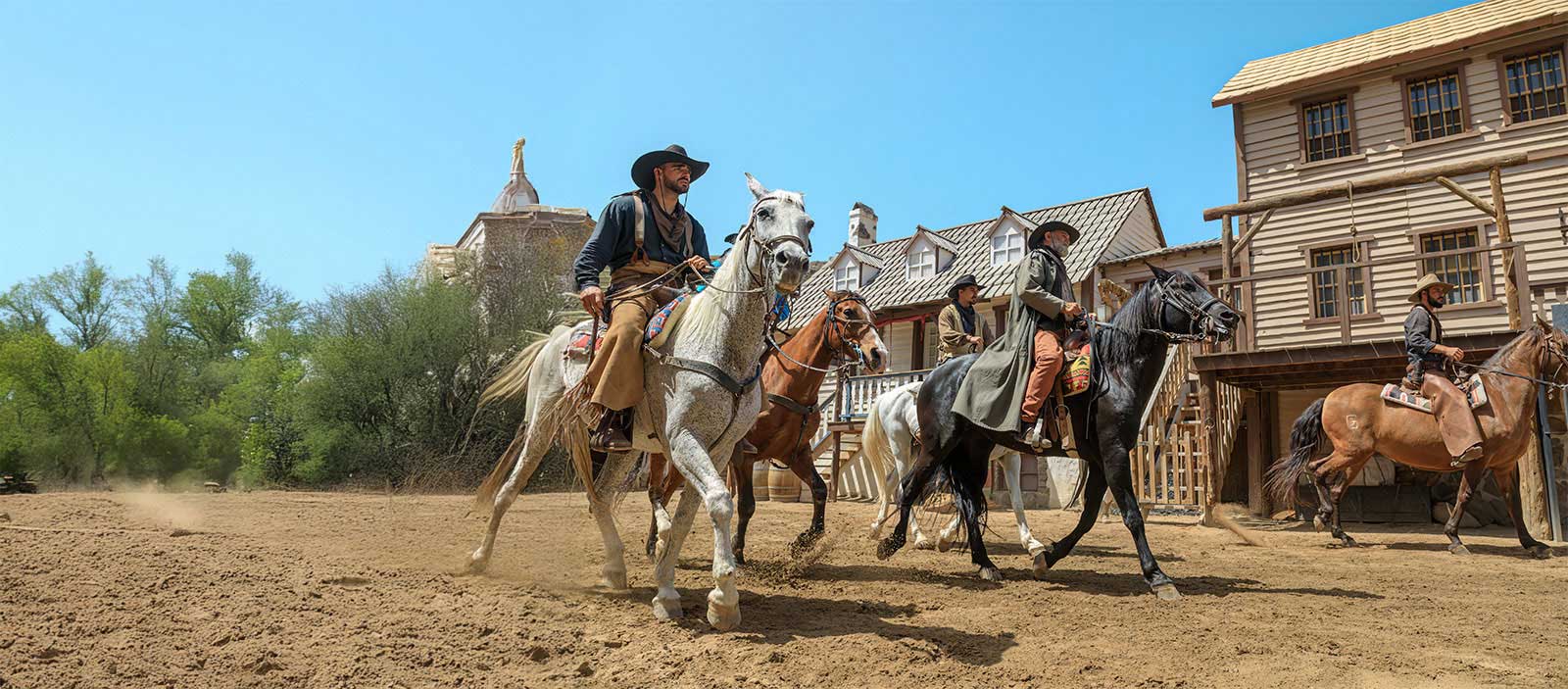 A group of four horse riders in Western attire ride through a sandy area, with rustic wooden buildings and greenery in the background.
