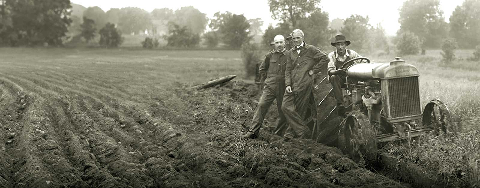 Three men stand in a field, one driving a small tractor.