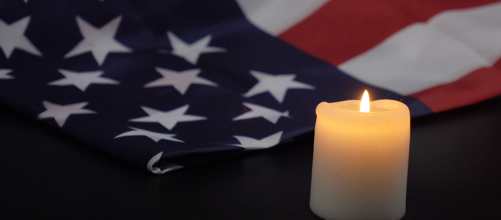 A lit white candle sits in front of a folded American flag, with red and white stripes and a blue field of white stars, against a dark background.