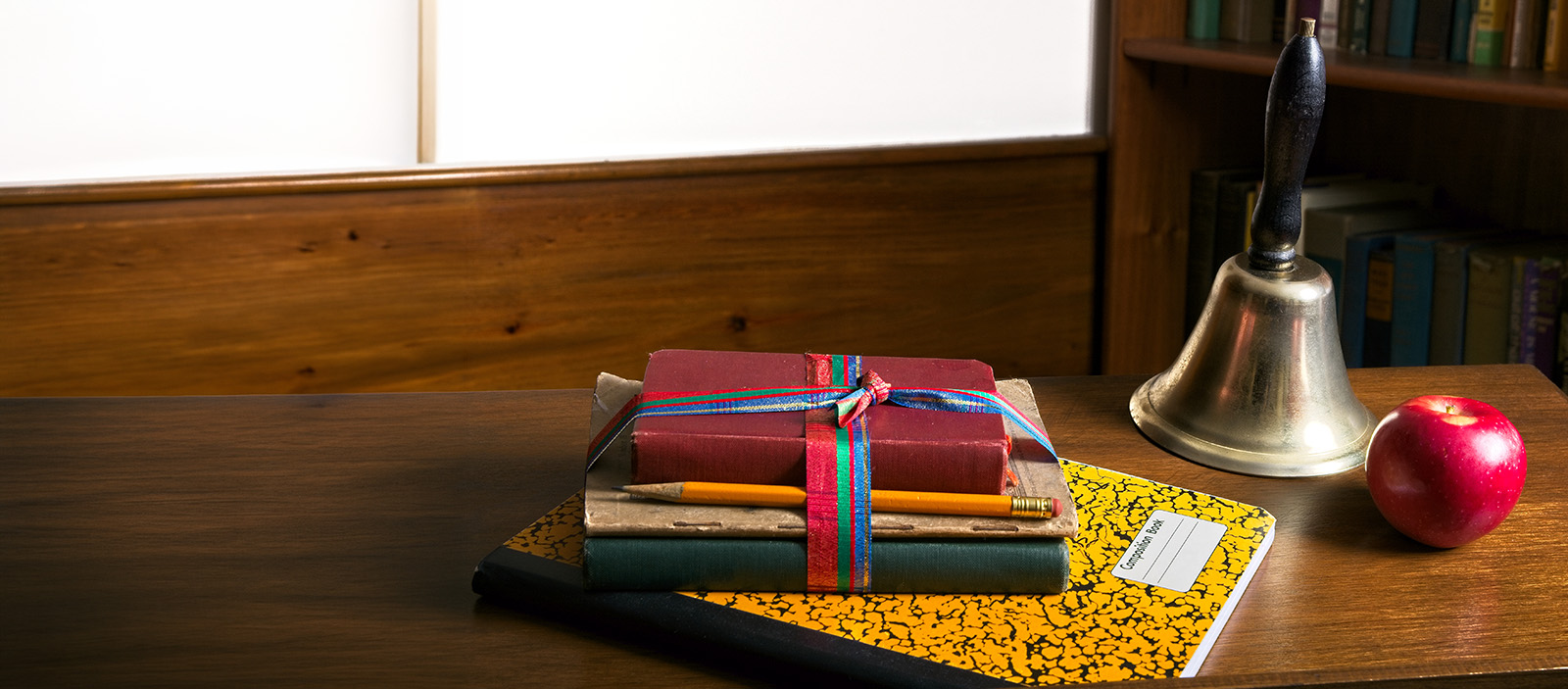 A wooden desk featuring stacked books wrapped in ribbon, a yellow pencil, a composition notebook, a brass bell, and a red apple.