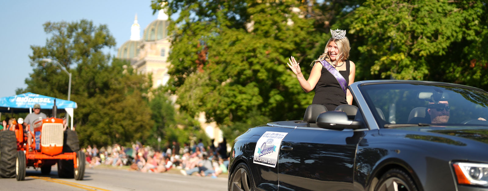 A young woman in a black dress, wearing a sash and crown waves to the crowd.