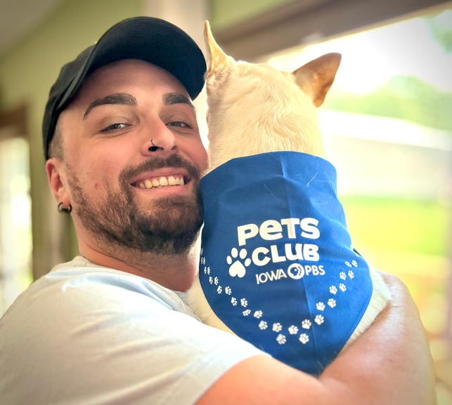 A man holds a small dog who is wearing an Iowa PBS Pets Club bandana.