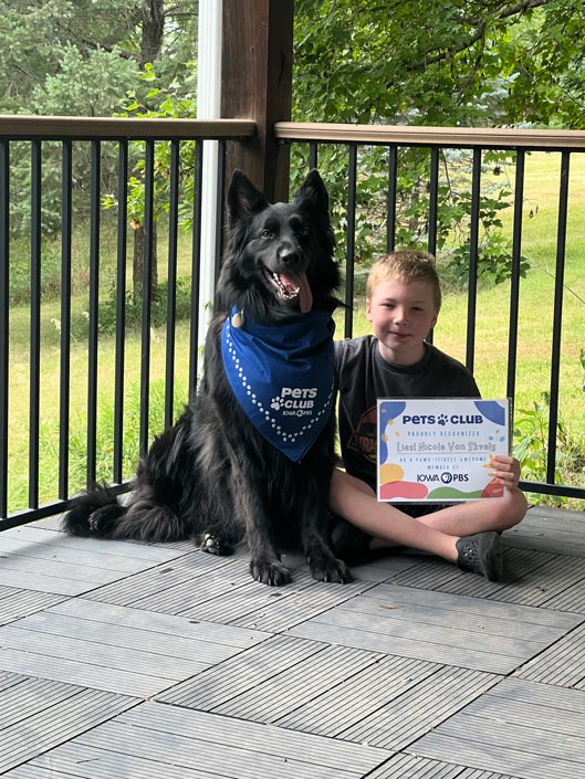 A large black dog sits next to a young boy holding an Iowa PBS Pets Club certificate.