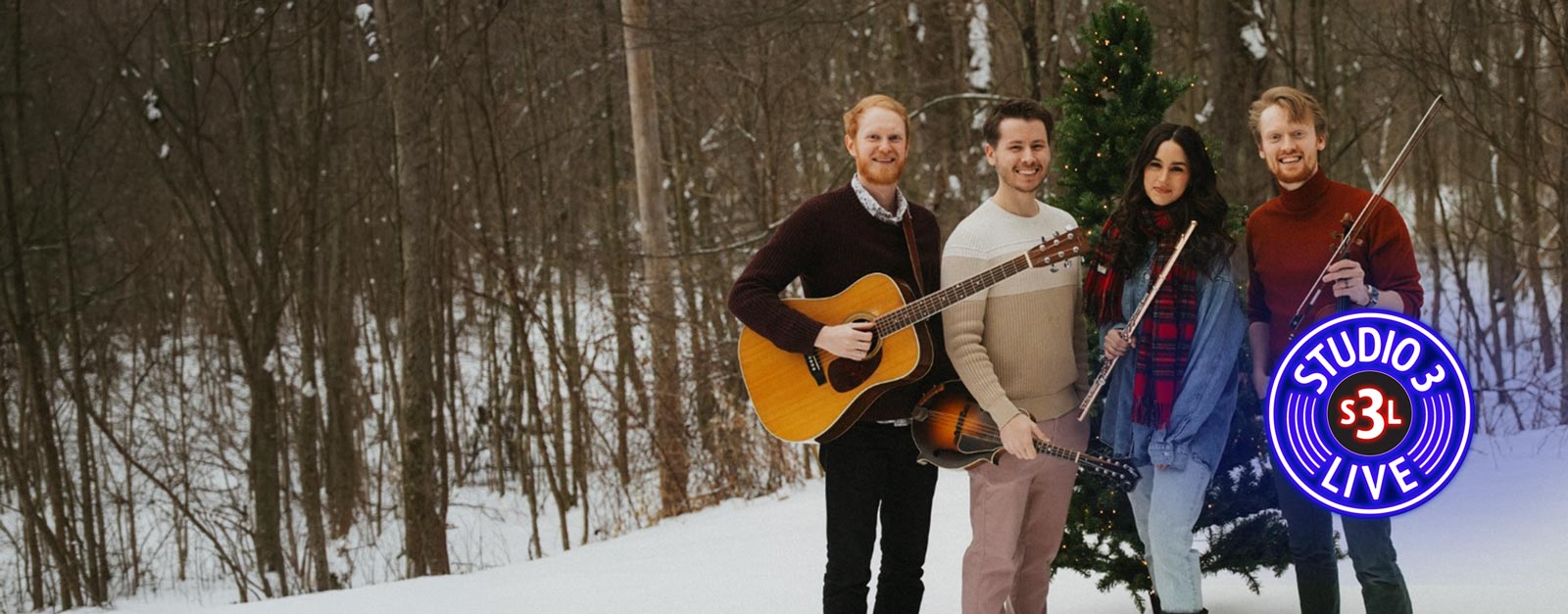 Three men and one woman dressed in winter gear stand on the edge of a snow covered forest.