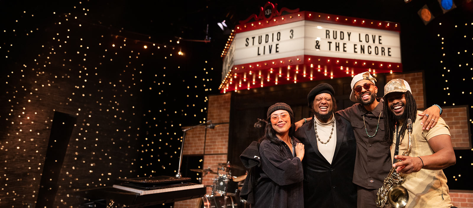 Four musicians stand under the Studio 3 Live marquee.