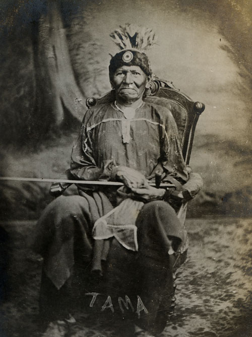 An older man dressed in traditional attire sits in a wooden chair with a feathered headdress and holds a long object.