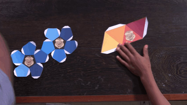 A dodecahedron in shades of blue with the Odd Squad gold shield in the middle cut out ready to be folded to form a 3 dimensional dodecahedron and an image of a tetrahedron with each side a different color from yellow, light orange, orange and red with the gold Odd Squad shield on the orange triangle. Ready to be folded into a three-dimensional tetrahedron.