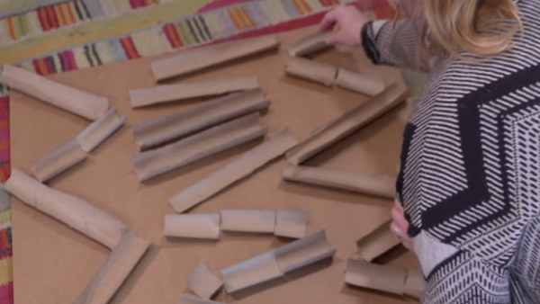 A woman laying out paper towel rolls along the lines on the cardboard. The paper towel rolls have been cut in half to allow a marble to roll along the inside of the tube.