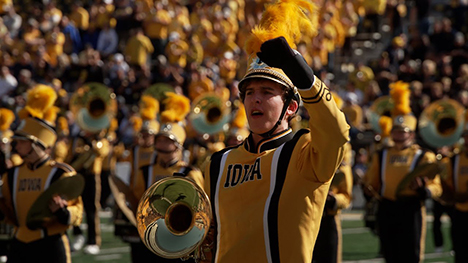 University of Iowa Marching Band member singing with fist in the air and instrument under other arm.