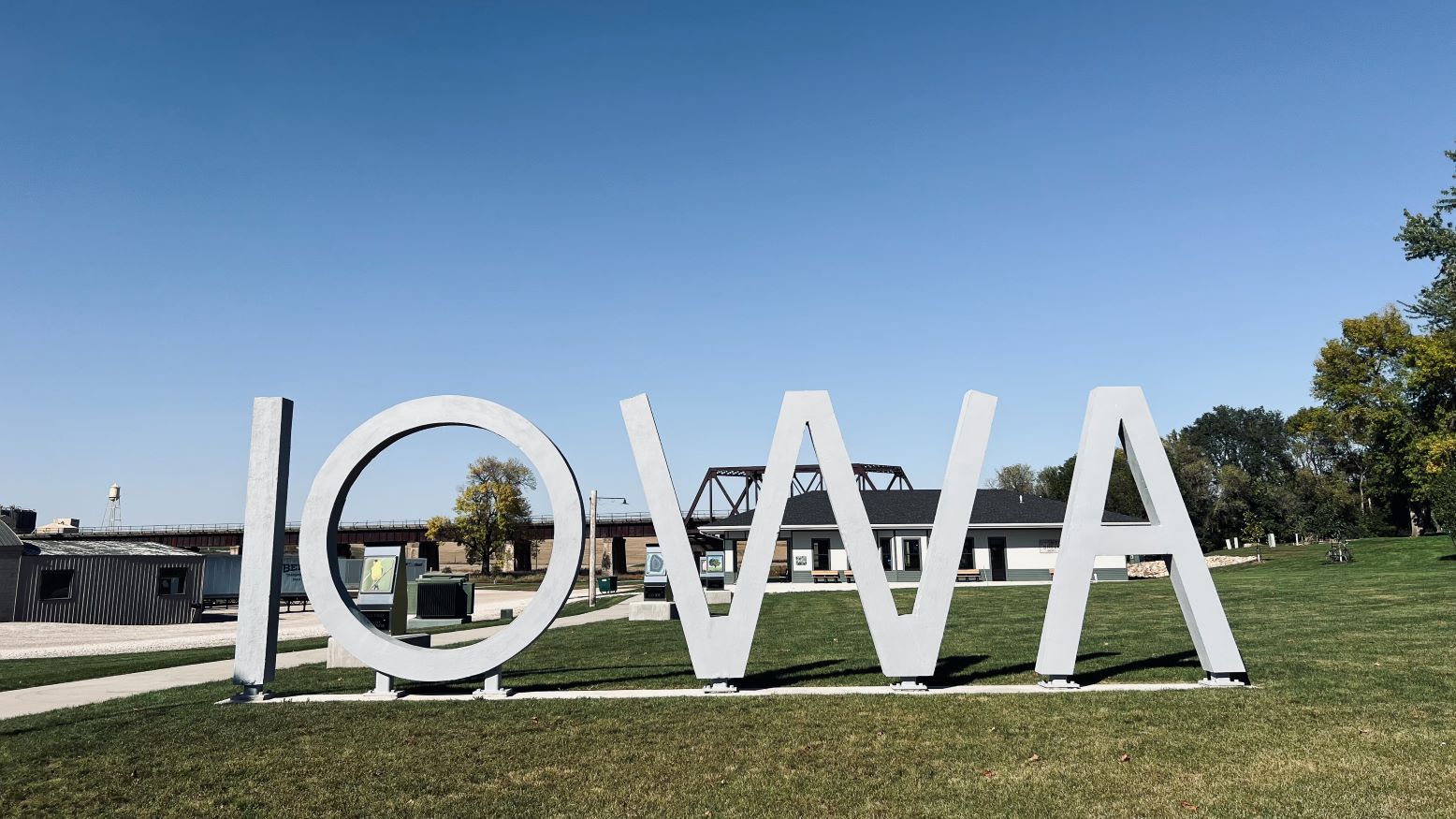 A sculpture of the word "IOWA" in large, white, capital letters. The sculpture stands on grass and there are buildings and a bridges in the background, showing through behind the letters. 