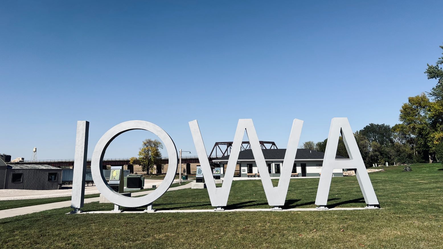 A sculpture of the word "IOWA" in large, white, capital letters. The sculpture stands on grass and there are buildings and a bridges in the background, showing through behind the letters.