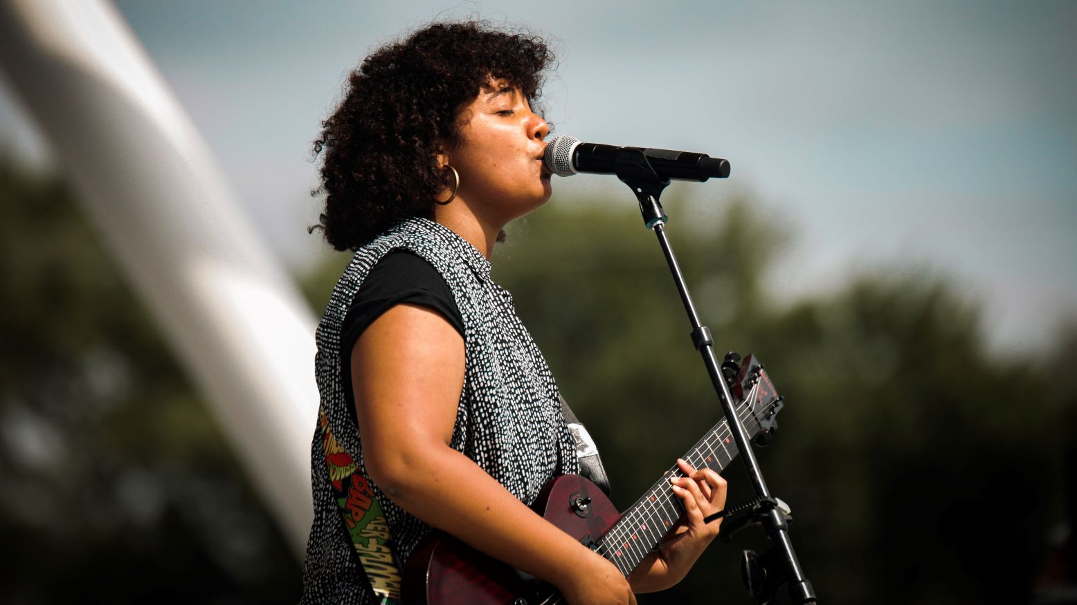 Musician Charlotte Blu, a young Black woman, plays guitar and sings into a microphone as she performs at an outdoor venue. 