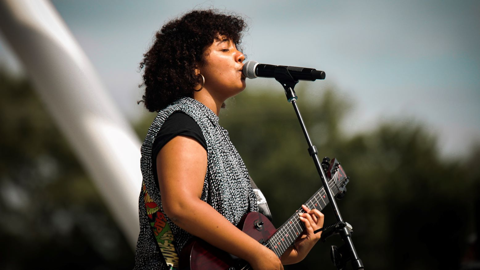 Musician Charlotte Blu, a young Black woman, plays guitar and sings into a microphone as she performs at an outdoor venue.