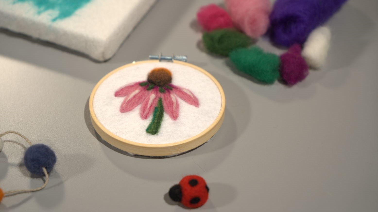 A few needle felting crafts and supplies on a gray surface. In the center is a circular craft made in an embroidery hoop depicting a flower with pink petals using felt. Below that is a small ladybug craft made from felt. There are piles of felt fibers in the top right corner of the image, and in the top left corner is part of a canvas. 