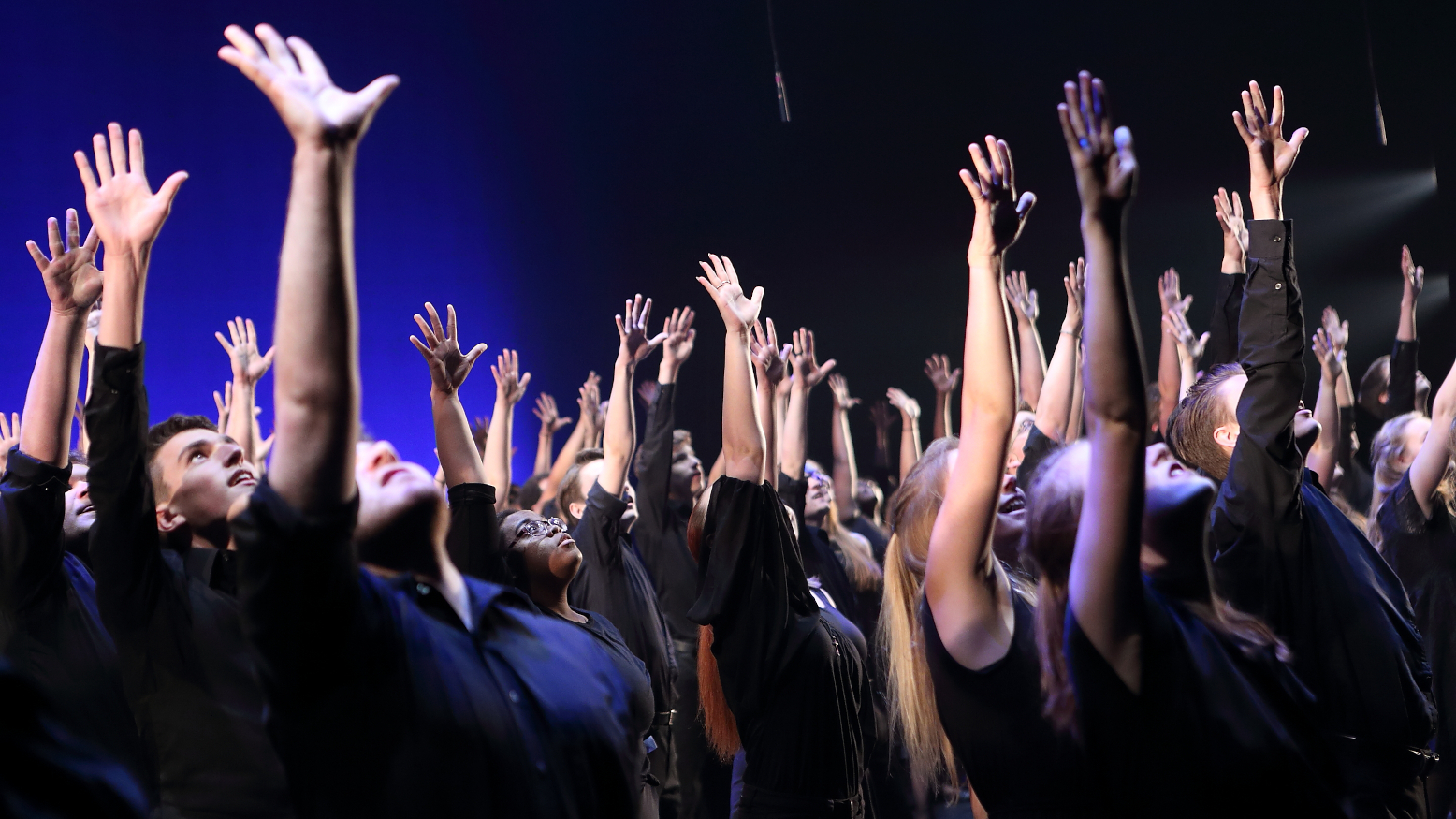 High school students on stage, each one with one arm raised in the air and looking toward the ceiling.