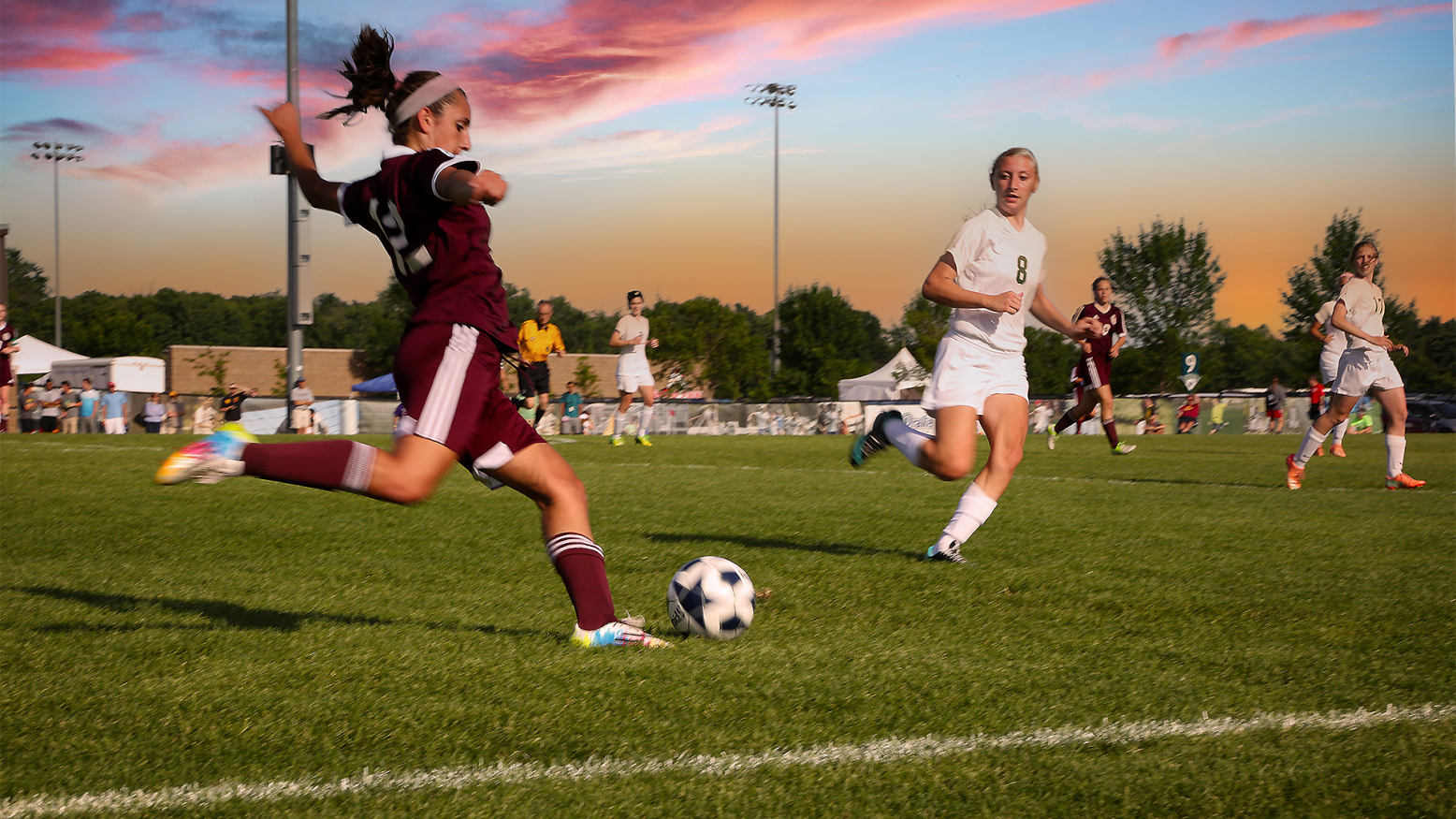 A soccer player in a maroon uniform is in mid-kick of a soccer ball. Another player wearing a white uniform approaches her to defend the ball from advancing down the field. Additional players are in the background, wearing maroon or white uniforms, as well as a referee wearing a yellow uniform. 