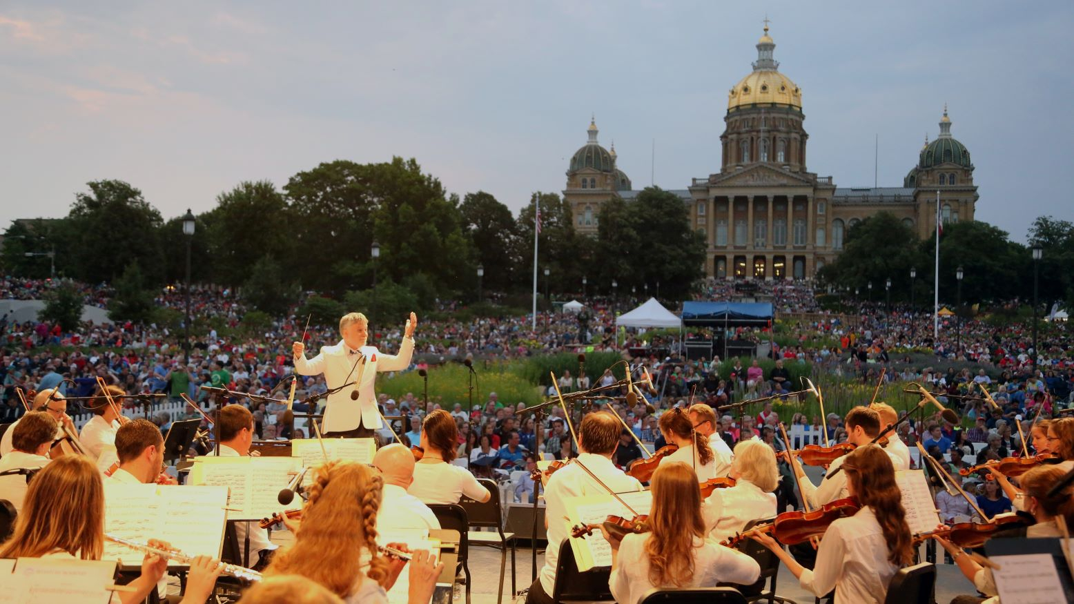 In the foreground, the Des Moines Symphony wearing white shirts plays at Yankee Doodle Pops. The Iowa Capitol and a crowd on its lawn are in the background.