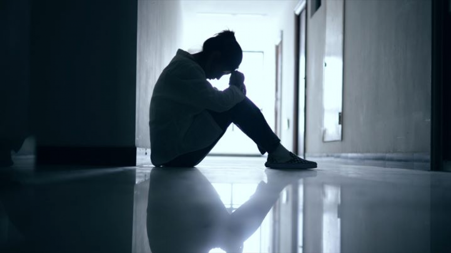 A woman sits on a slightly reflective floor of an empty hallway. Her knees are pulled up toward her chest and she rests her hands on her knees. Her forehead is resting on her hands. The woman is backlit, so few details are visible other than her silhouette. 