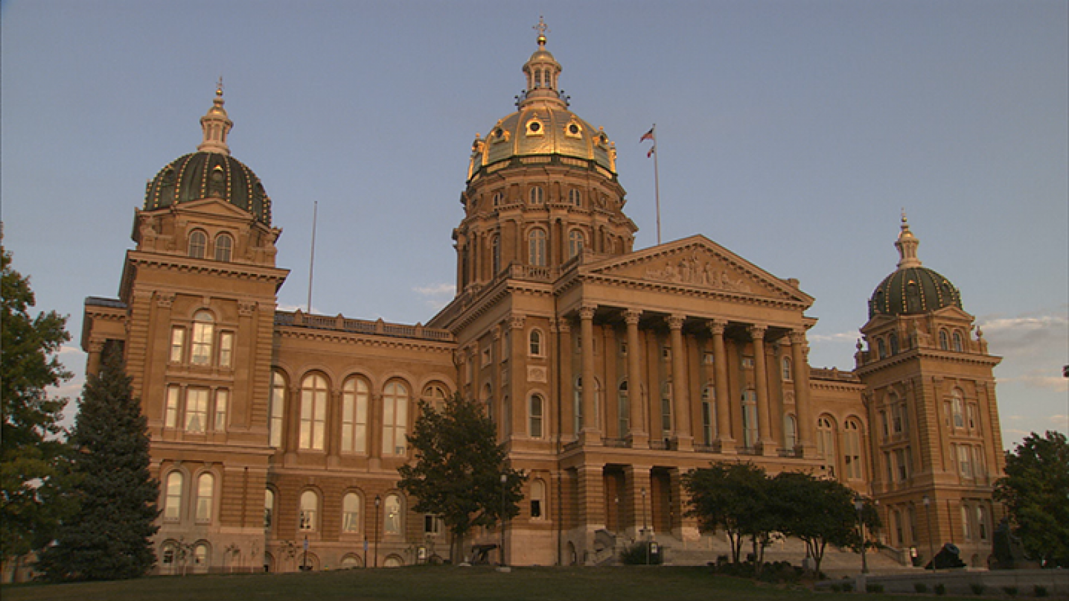 An image of the Iowa State Capitol taken from the lawn. A few trees stand in front of the building, which features multiple domes. The largest dome is in the center and is gold. 