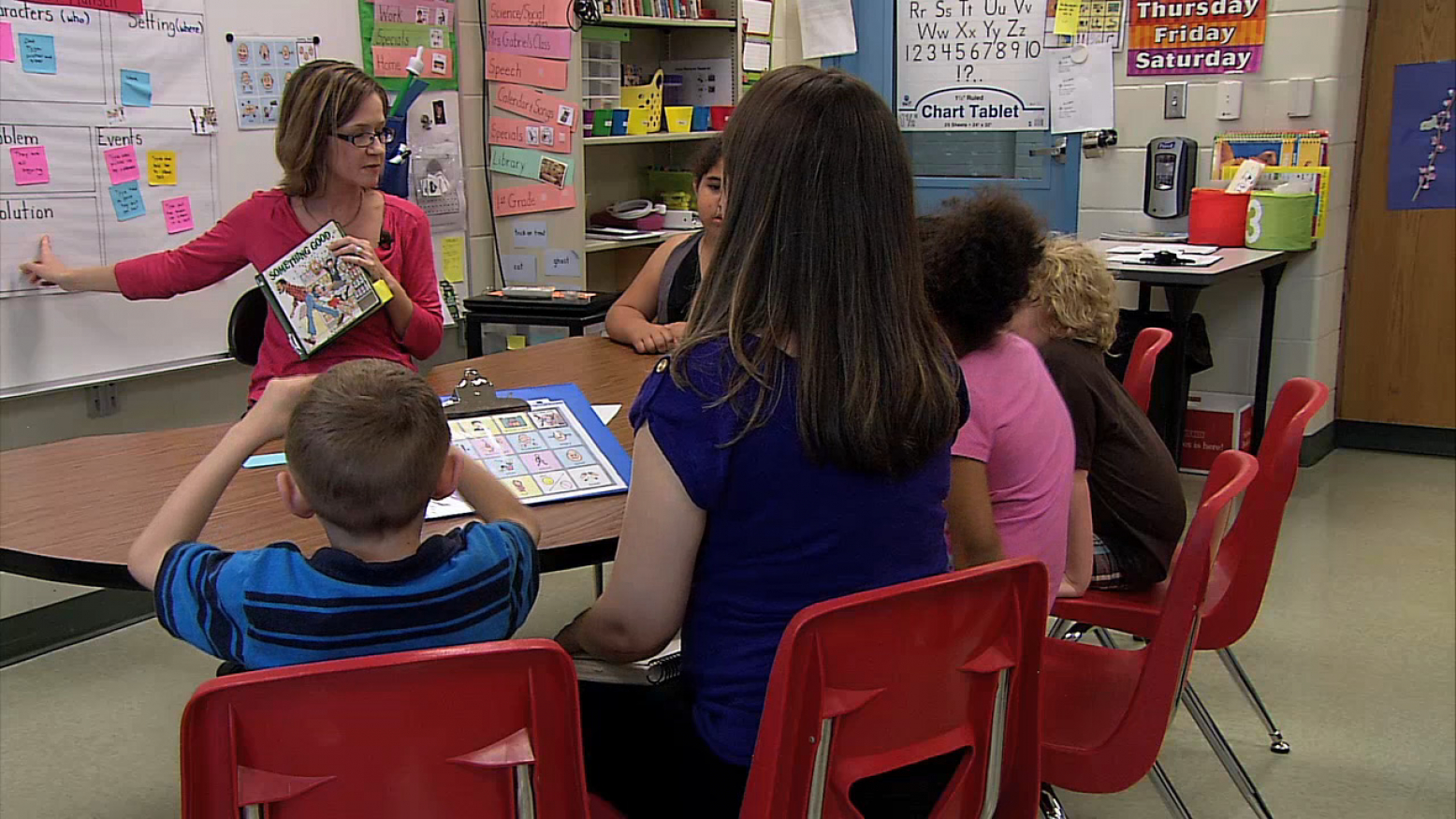 Elementary students sit at a table as their teacher points at the white board.