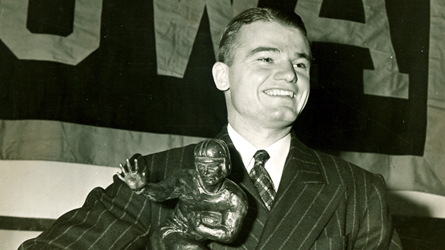 A black and white image of a young, smiling Nile Kinnick holding the Heisman Trophy. In the background, part of a flag that reads "IOWA" is visible. 