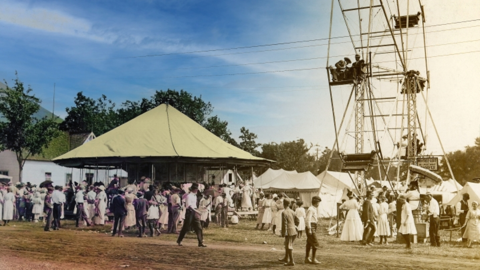 A semi-colorized archival image of fairgoers at the Iowa State Fair. People of all ages are standing and walking around and through tents. A ferris wheel is in the background of the right side of the image.