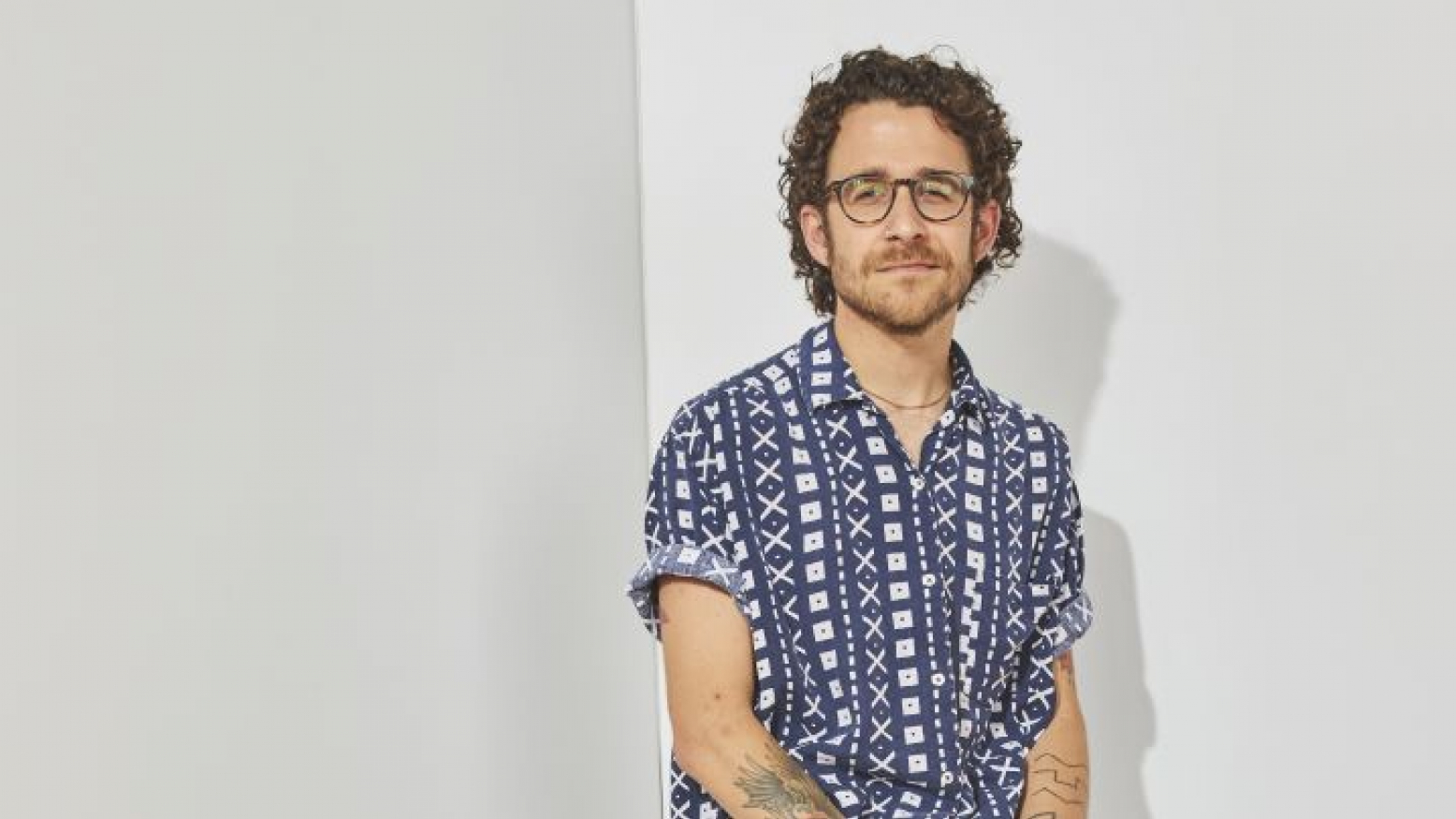 An image of Mickey Davis, a white man with short, curly brown hair wearing glasses. He is in front of a white wall and is wearing a short sleeved button-down shirt with a blue and white geometric pattern.