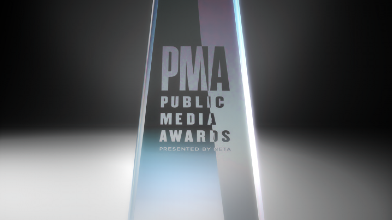 An image of a Public Media Awards trophy. The trophy is made of glass and is a pyramidal shape. Etched in the glass is the logo for the awards, reading "PMA Public Media Awards Presented by NETA."