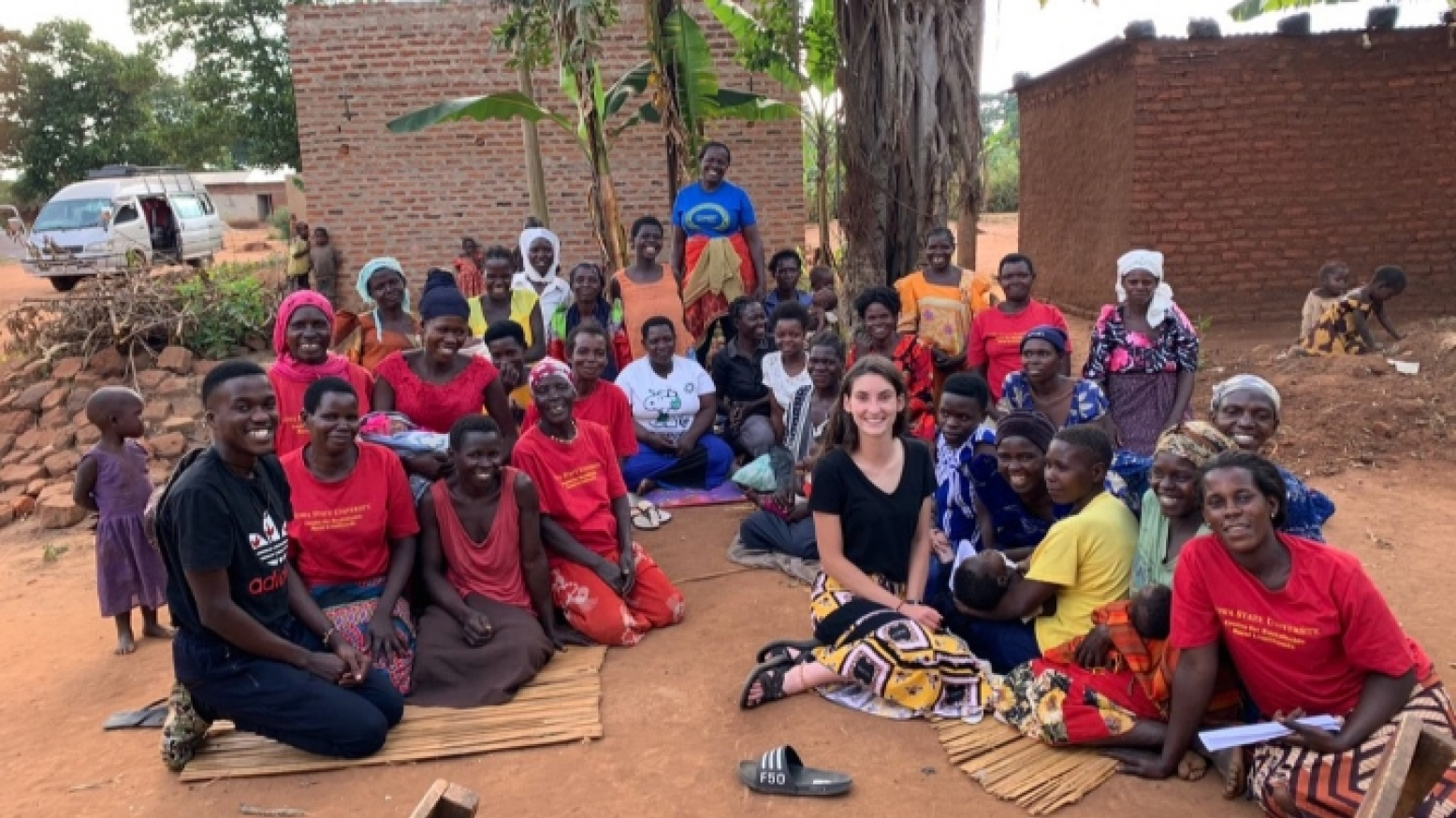 Sofia Fernandez, a young white woman with brown hair, sits on the ground surrounded by approximately 30 Ugandan people. All are smiling for a photo.