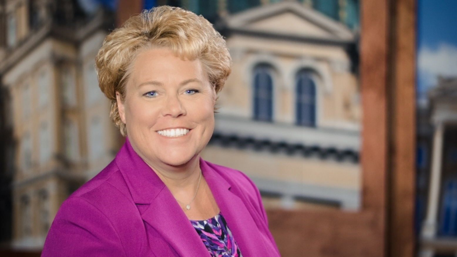 An image of Molly Phillips, a white woman with short blonde hair wearing a magenta blazer, standing in front of the Iowa Press television set.