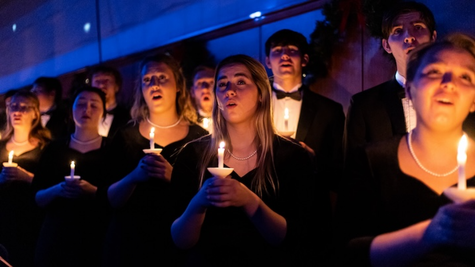 Wartburg choir members dressed in tuxedos and black dresses hold lit candles as they sing. 