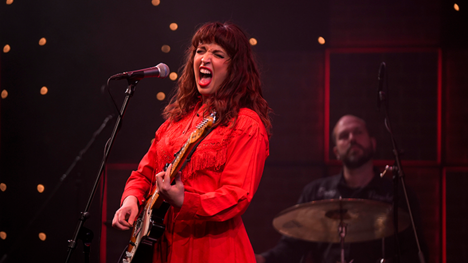 An image of Elizabeth Moen performing on the Studio 3 LIVE set. She has long red hair, is wearing a red long-sleeved dress and is playing a guitar while singing into a microphone. In the background, a bald man with a beard is seated playing the drums.