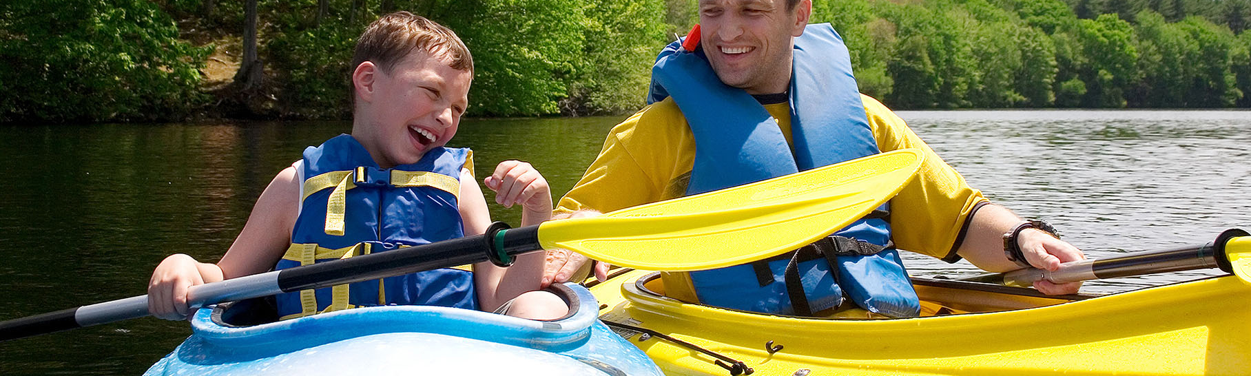 A boy and a man smiling while sitting in kayaks on a lake.