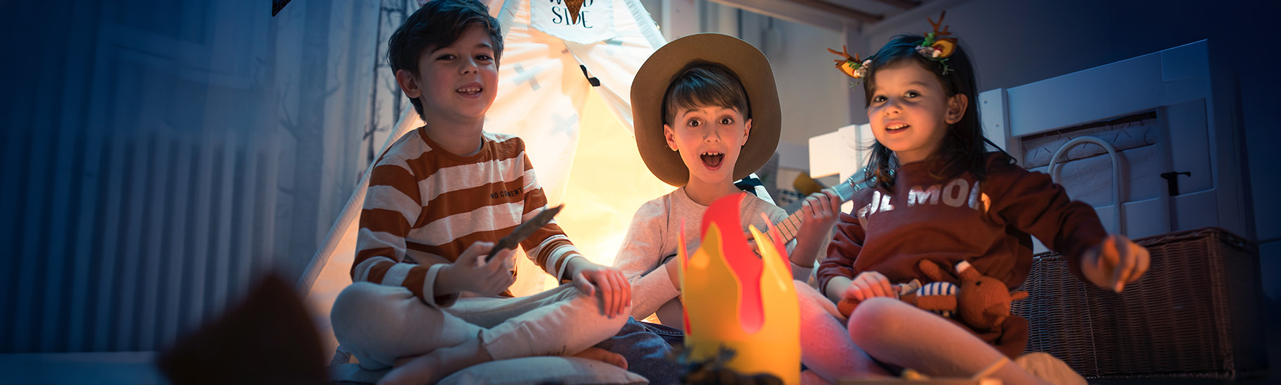Three children sitting around a faux campfire in a cozy indoor setting, smiling and enjoying playtime together.