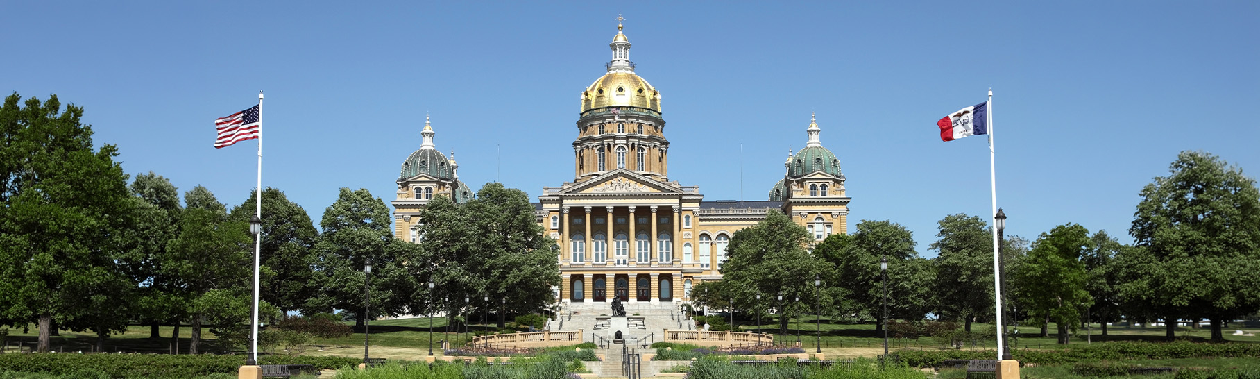 A historic government building with a golden dome and classical architecture, surrounded by trees and featuring the American and Iowa flags on either side.